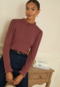Prugasti dugih rukava top u tamnocrvenoj i krem boji s volanima na izrezu i naglašenim rukavima, u kombinaciji s tamnoplavim trapericama visokog struka i smešnim remenom.