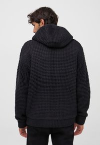 Czarny dzianinowy, oversized sweter z kapturem o fakturowanym wzorze, z prążkowanymi mankietami i dołem, prezentujący swobodny design z widoku z tyłu.