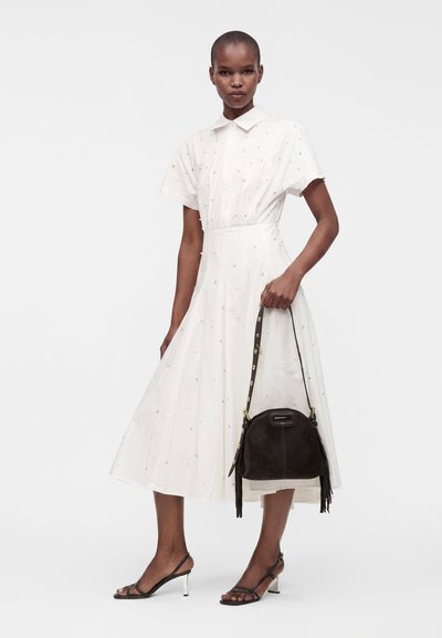 Femme portant une robe midi blanche à manches courtes avec de petits pois, des sandales noires à talons et tenant un sac à main noir avec des franges.
