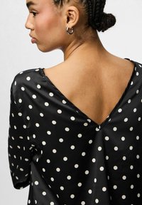 Blusa de satén negro con lunares blancos, escote en la espalda en forma de V y mangas cortas. Textura suave y corte relajado.