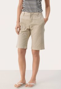 Pantaloni corti in cotone beige con taglio dritto, tasche laterali e orlo risvoltato, abbinati a una canotta a righe bianca e nera e sandali.