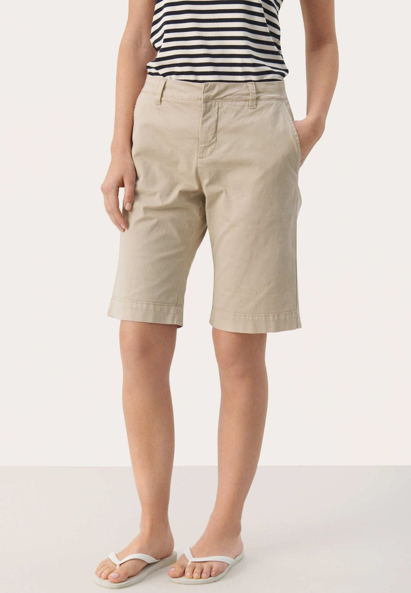 Pantaloni corti in cotone beige con taglio dritto, tasche laterali e orlo risvoltato, abbinati a una canotta a righe bianca e nera e sandali.