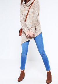 Beżowy sweter z golfem o teksturowanym splotem, zestawiony z niebieskimi obcisłymi dżinsami i brązowymi botkami za kostkę. Do tego mała brązowa torebka na ramię.