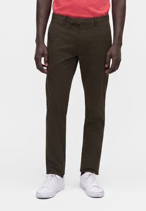 Homme portant un pantalon marron foncé fuselé, une chemise rouge rentrée et des baskets blanches, debout devant un fond clair uni.