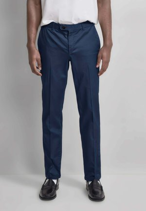 Persona che indossa pantaloni slim blu navy, mocassini neri e una camicia bianca infilata, in piedi davanti a uno sfondo grigio chiaro uniforme.