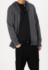 Persona vestida con una sudadera de cremallera gris oscuro sobre una camiseta negra y pantalones negros, con una mano en el bolsillo y gafas de sol colgando del cuello de la camiseta.