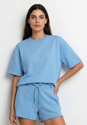 Vrouw met lang donker haar, gekleed in een effen lichtblauw oversized T-shirt met korte mouwen en bijpassende korte broek met trekkoord, staand tegen een witte muur.