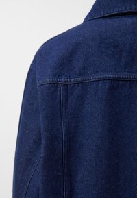 Tummansininen farkkutakki, jossa on klassinen kaulus, näkyvät saumat ja teksturoitu pinta, esillä takakuvasta ja hartia-alueelta.