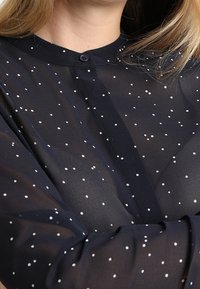 Mörkblå genomskinlig blus med små vita prickar, rund krage och knappar i full längd. Lätt, delikat tyg med en slät textur.