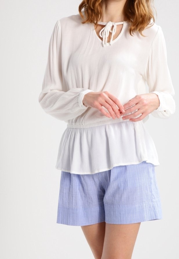 Femme portant un chemisier blanc à manches longues avec un col à nouer et un short rayé bleu et blanc, debout les mains jointes.