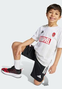 White short-sleeve t-shirt with Spider-Man logo and patterned design, paired with black shorts featuring white accents. Black and red shoes.