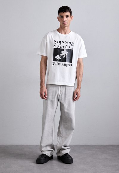 T-shirt blanc à manches courtes avec impression graphique noire ; coupe décontractée. Pantalons en denim gris clair ; chaussures noires texturées. Design simpliste.