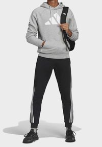Sudadera gris con logo blanco, pantalones de chándal negros ajustados con rayas blancas y zapatillas deportivas negras. El modelo sostiene una mochila negra.