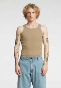 Tan ribbet tanktopp med brede skulderstropper, kombinert med lyse denimshorts. Merkbare tatoveringer synlige på begge armer.
