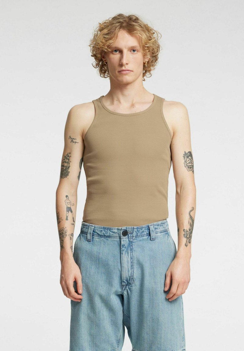 Tan ribbet tanktopp med brede skulderstropper, kombinert med lyse denimshorts. Merkbare tatoveringer synlige på begge armer.