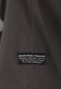 Tela negra con una textura acanalada que presenta una etiqueta prominente con el texto "ESCAPA DE / LO DESCONOCIDO", en blanco y con forma rectangular.