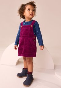 Vestito di velluto a coste di colore rosa intenso con ampia tasca frontale e bottoni in ottone. Maglia a maniche lunghe blu con pois bianchi. Stivaletti alla caviglia blu scuro.