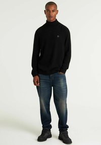 Fekete garbó pulóver, puha anyagból, kék farmernadrággal, melynek elhalványult mintája van. A modellen fekete túracipők vannak.