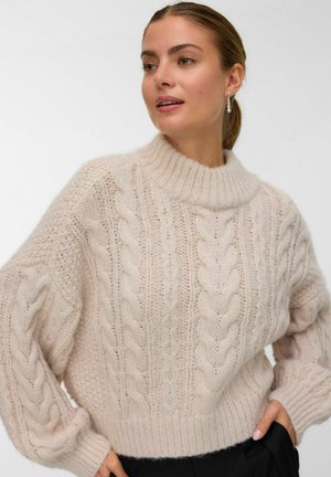 Vrouw in een crème-kleurige chunky kabelgebreide trui met een hoge ronde hals en lange pofmouwen, staande met een hand in de zak van een zwarte broek.