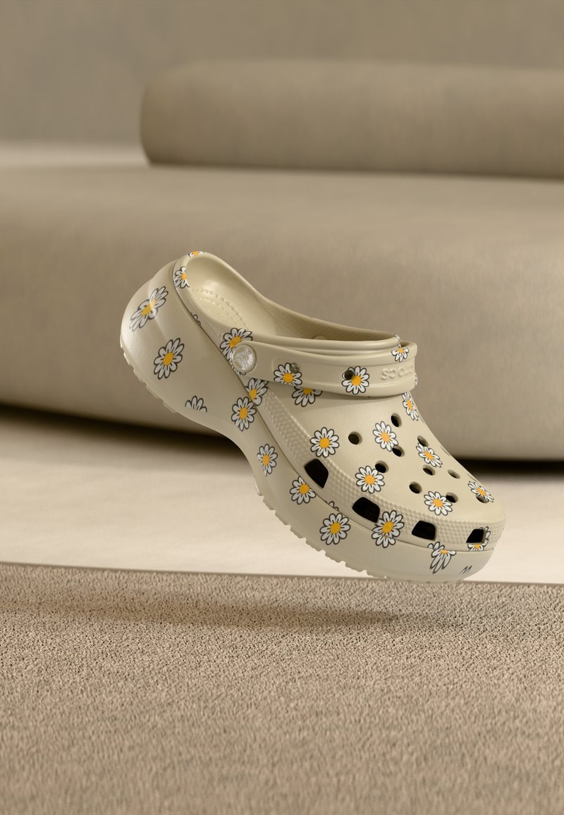 Μπεζ κλογκ με μοτίβα μαργαρίτας να αιωρείται πάνω από μοκέτα σε ένα δωμάτιο με ουδέτερους τόνους και καναπέ στο φόντο.