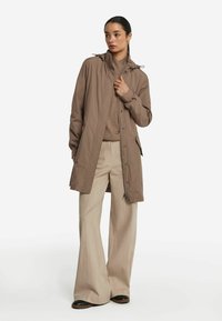 Ljusbrun vattentät jacka med dragsko på huven, dragkedja fram och sidofickor, kombinerad med beiga vida byxor och svarta skor.