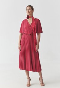 Abito rosso plissettato con maniche corte e scollo a V, arricchito da una cintura di tessuto e un accento floreale. Indossato con sandali beige con tacco alto.