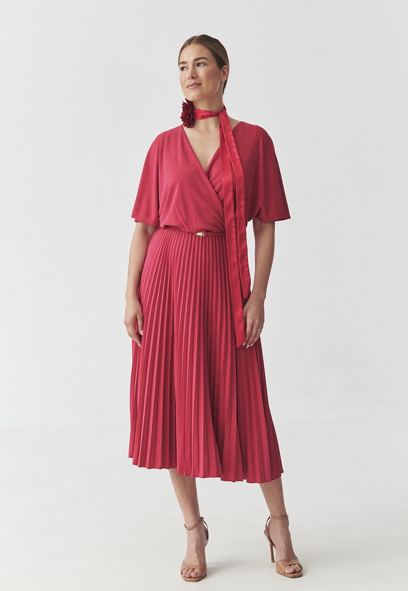 Abito rosso plissettato con maniche corte e scollo a V, arricchito da una cintura di tessuto e un accento floreale. Indossato con sandali beige con tacco alto.