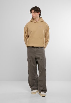 Haut de survêtement beige surdimensionné avec une poche kangourou à l'avant, associé à un pantalon cargo gris foncé avec plusieurs poches et une coupe décontractée.