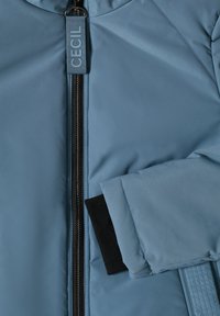 Manica e parte anteriore di giacca imbottita blu con zip nera e linguetta in tessuto etichettata "CECIL", che mostra il polsino interno nero.