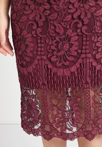 Burgundská krajková sukně se květinovými vzory a volánkovým lemem, vrstvená s průsvitnou tkaninou. Texturovaný design s jemnými detaily.
