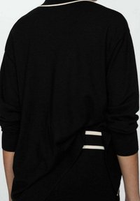 Černý oversized svetr s elegantním designem, který má jemnou texturu, žebrované manžety a kontrastní bílé pruhy na lemu a na zádě.