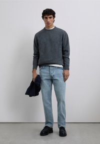 Suéter de punto gris, camiseta blanca, jeans azul claro, sosteniendo una chaqueta azul marino, zapatos negros, de pie contra un fondo claro y liso.