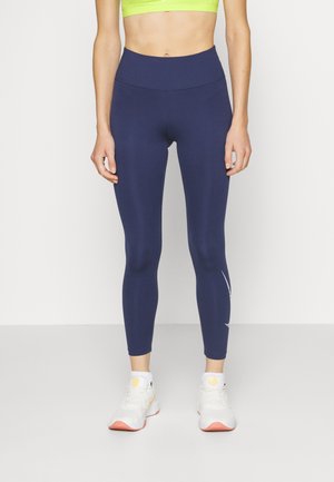 Leggings taille haute en marine, fabriqués en tissu extensible à la texture lisse. Présente un détail de design blanc sur la partie inférieure de la jambe. Associés à des baskets blanches.