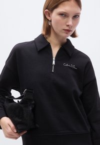 Felpa nera con collo con zip e logo ricamato, abbinata a una piccola borsa nera testurizzata con manico.