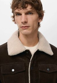 Brun kordfløjlsjakke med beige shearling-krave, med to brystlommer og trykknaplukning. Båret over en cremefarvet sweater.