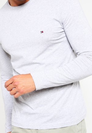 Hombre vistiendo una camisa de manga larga gris claro con un pequeño logo rectangular rojo, blanco y azul marino en el pecho, con pantalones beiges parcialmente visibles.