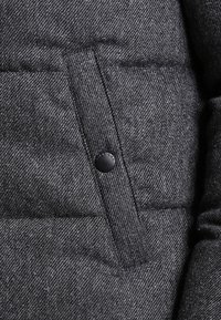 Segmento de chaqueta acolchada gris con un tejido texturizado, que presenta un botón a presión negro y un detalle de bolsillo visible.
