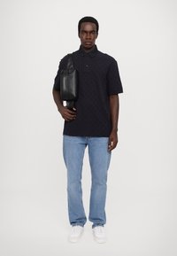 Navy polo skjorte med teksturert mønster, svart crossover-veske, blå jeans og hvite sneakers. Modellen står mot en ensfarget bakgrunn.
