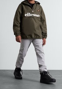 Olijfgroene pullover jas met een voorktas, wit "ellesse" logo en zwarte accenten, gecombineerd met lichtgrijze cargobroek en zwarte sneakers.