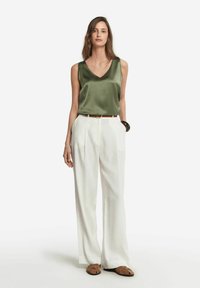 Oliwkowa, satynowa, bezrękawnikowa bluzka z dekoltem w szpic, w połączeniu z białymi, wysokimi spodniami o szerokich nogawkach oraz brązowymi, wsuwanymi butami.