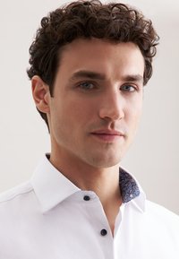 Man met krullend donker haar, gekleed in een wit overhemd met een geprint binnenkraag, tegen een lichte achtergrond.