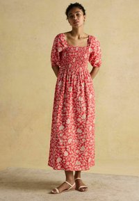 Rotes Maxikleid mit quadratischem Ausschnitt, gerafftem Oberteil, puffärmeligen kurzen Ärmeln und einem fließenden Rock. Getragen mit neutralen Sandalen.