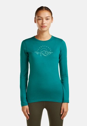 Młoda kobieta w turkusowej koszulce z długim rękawem z grafiką gór i napisem „ICEBREAKER”, w ciemnooliwkowych spodniach, stojąca na białym tle.