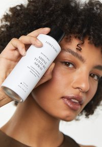 Vaporisateur de brillance dans une bouteille blanche mate avec un capuchon noir. L'étiquette présente le nom du produit et ses avantages en texte noir. Tenue à la main contre la peau.