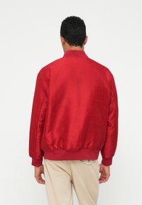 Punane pommitaja jope, millel on sile satiinviimistlus, soonikkoes varrukad ja alläär ning peen reljeefne muster. Tagapoolne vaade, kantud beežide pükstega.