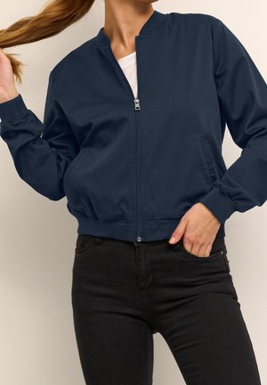 Femme portant une veste bomber bleu marine zippée sur une chemise blanche et un jean noir, tenant ses cheveux d'une main et l'autre main dans la poche.