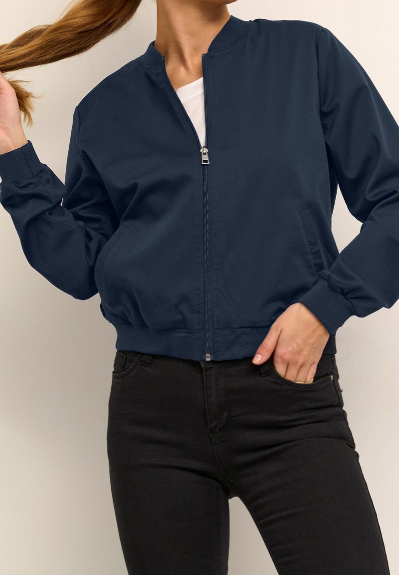 Femme portant une veste bomber bleu marine zippée sur une chemise blanche et un jean noir, tenant ses cheveux d'une main et l'autre main dans la poche.