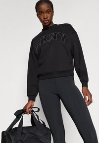 Czarna bluza z logo DKNY w teksturowanym wzorze. Połączona z czarnymi legginsami, trzyma czarną torbę sportową z podwójnymi uchwytami.