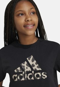 Camiseta de algodón negra con un gran logotipo de Adidas en estampado de leopardo beige en el frente. Cuello redondo, mangas cortas y textura suave.