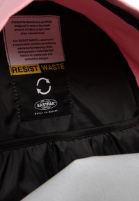 Interior de tela negra de una mochila Eastpak que presenta una etiqueta RESIST WASTE en gris con texto negro y amarillo, diseñada para la sostenibilidad.